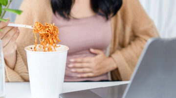 Woman eats noodles at desk, holds stomach