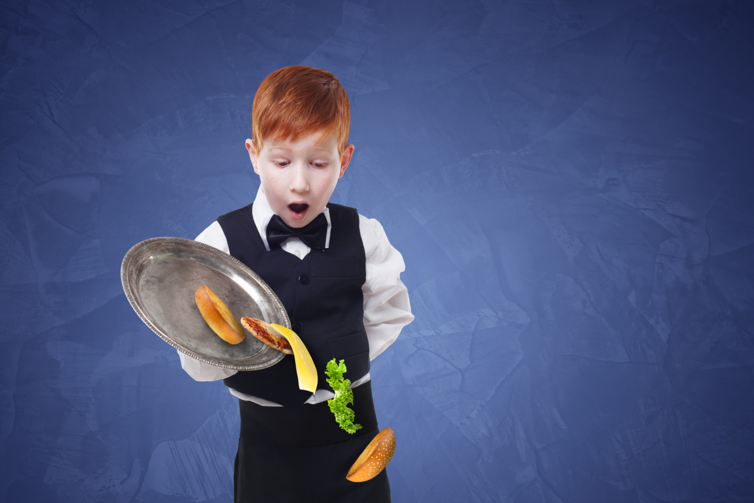 Clumsy little waiter drops food from tray serving hamburger. Cheeseburger falling with separated toppings. Dropping burger layers. Redhead child boy in suit, failure at blue background