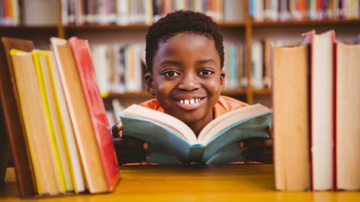 A Black boy reads a book and smiles while posing between other books at the library.