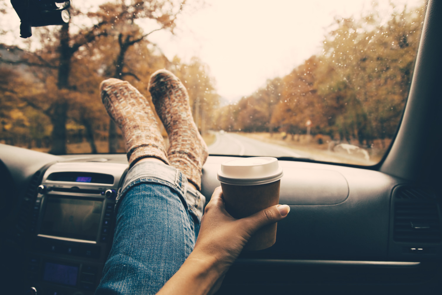 Woman feet in warm socks on car dashboard. Drinking take away coffee on road. Fall trip. Rain drops on windshield. Freedom travel concept. Autumn weekend. Filtered photo.