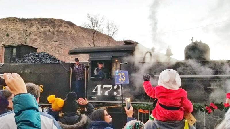 child in red holiday outfit sitting on their parent's shoulders waves at a vintage locomotive as it passes by
