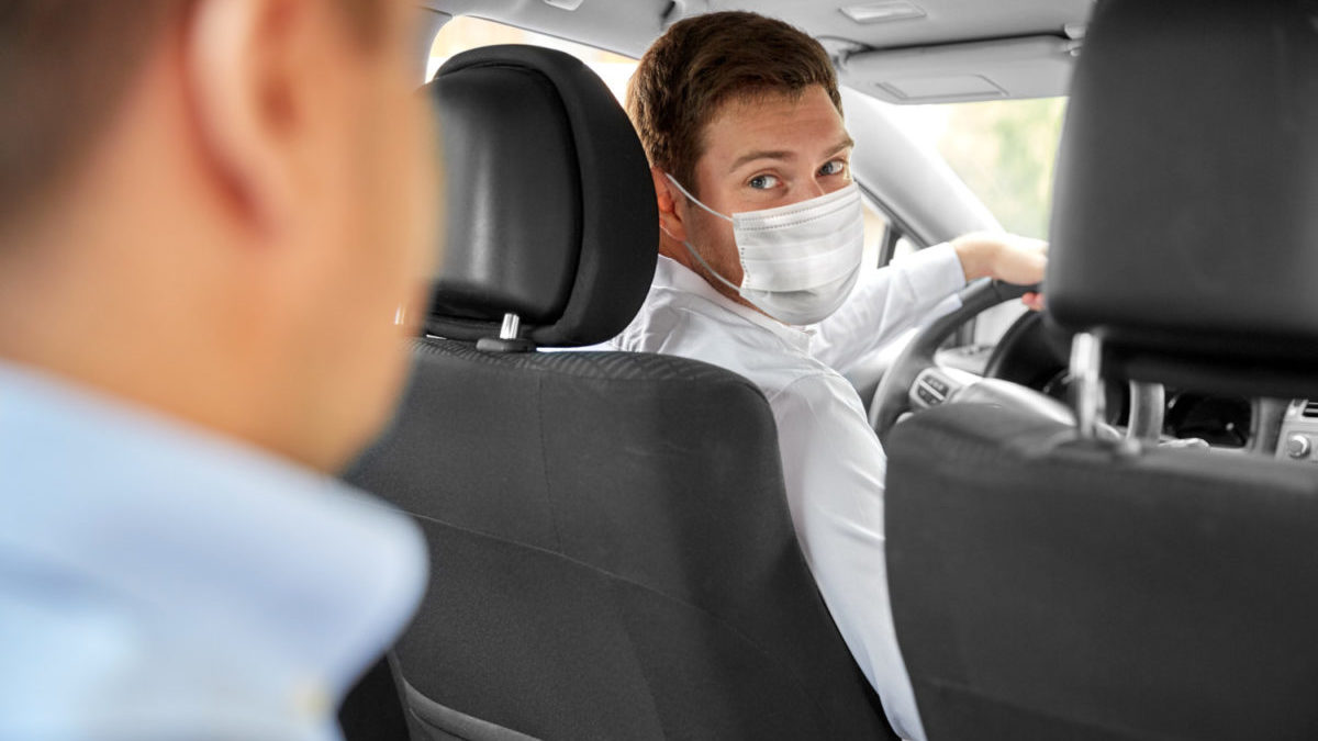 A ride-share driver looks back at his passenger while wearing a face mask.