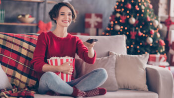 woman holding remote control and popcorn sitting on couch wearing red sweater