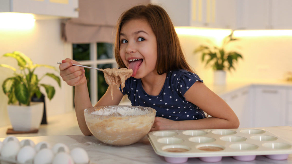 Cute little girl licking raw dough from spoon in kitchen.