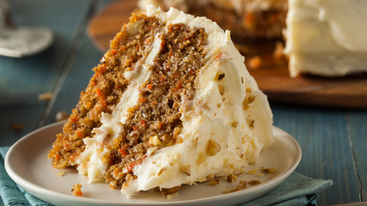 Slice of carrot cake with white frosting up close on plate with larger cake in background