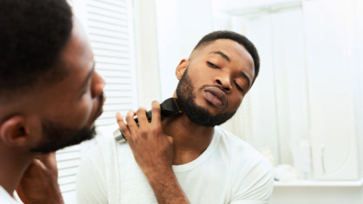 Young black man looking at mirror and shaving beard with trimmer.