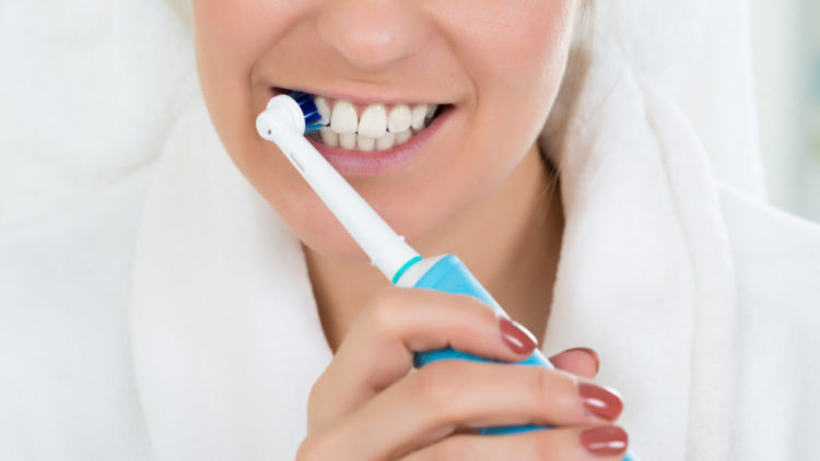 A white woman in a bathrobe brushes her teeth with an electric toothbrush.