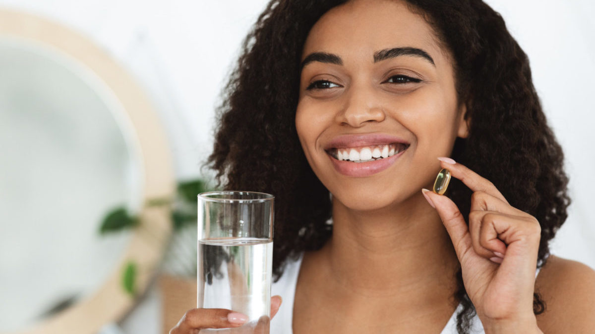 A young Black woman takes a fish oil supplement pill with water.