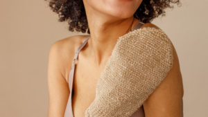 A Black woman uses an exfoliating mitt on her shoulder to remove dead skin.