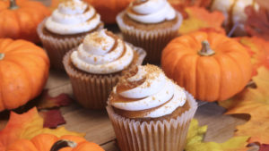 Frosted pumpkin cupcakes sit on a table with mini pumpkins and fall leaves.