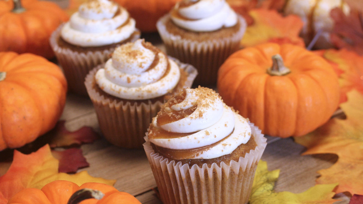 Frosted pumpkin cupcakes sit on a table with mini pumpkins and fall leaves.