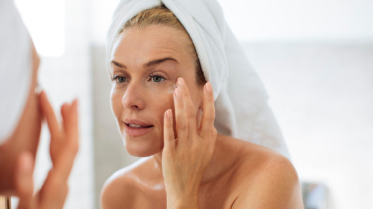 woman in towels looking in a mirror and putting face cream on