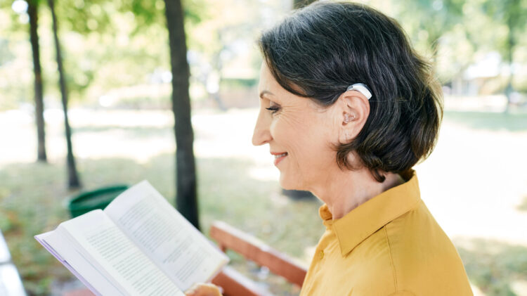 Mature woman with a hearing impairment uses a hearing aid in everyday life, reading a book in park, outdoor.