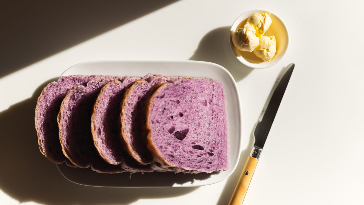 slices of purple-hued blueberry bread lying on a countertop next to a bread knife and small dish of butter