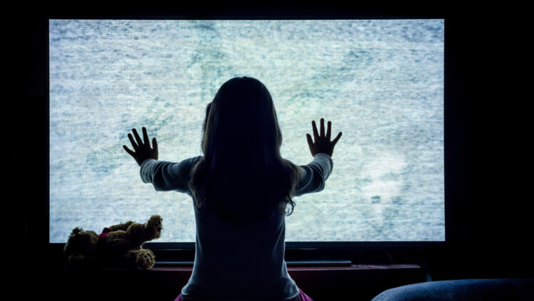 creepy image of young girl standing in the dark in front of a large, bright TV