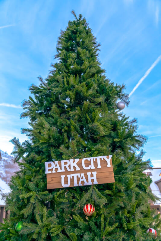 Christmas Tree with Park City Utah wooden sign. Outdoor Chistmas tree against snowy mountain and blue sky viewed on a sunny day. A Park City Utah wooden sign can be seen on the festive tree.