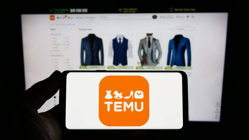 Photo of a person holding up a smartphone with Temu app open in front of a computer screen also with Temu website open