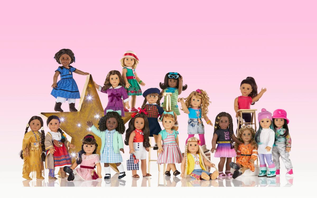 A collection of American Girl dolls in front of a pink backdrop