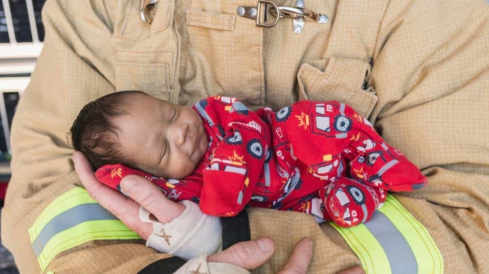 Newborn in a firefighter-themed onesie sleeps nestled in the arms of a firefighter
