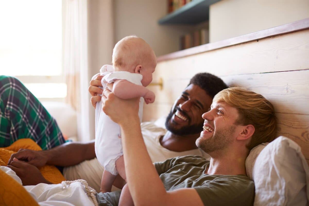 Loving Male Same Sex Couple Cuddling Baby Daughter In Bedroom At Home Together