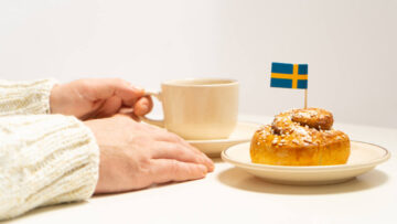 Swedish cinnamon bun with swedish flag on top and coffee cup in hand on white wooden table.