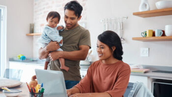 two parents in a kitchen with an infant, dad is holding baby and mom is working on computer