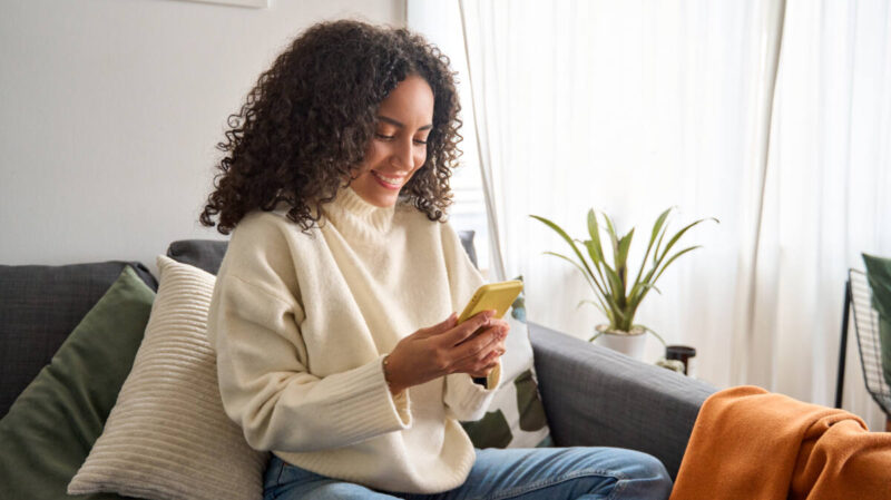 Woman sitting on couch, smiling as she uses smartphone
