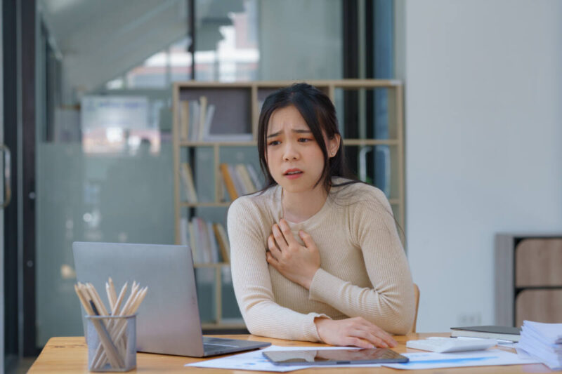 An Asian female has a heart attack pain, tension Sick employee feeling heartache while working on laptop at workplace at desk in office Sad woman in a suit keeps her hand on her chest indoor