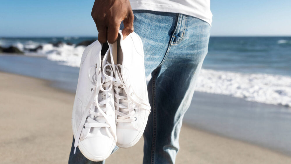 man holds pair of white sneakers on the beach